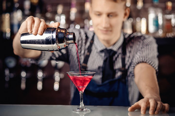 Expert barman making cocktail with shaker in martini glass, red. Dark background