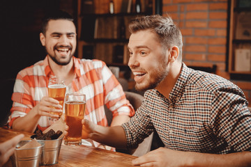 Friends young bearded men have fun in bar, clanging glasses of beer, laughing, on table they snacks cans that mimic buckets