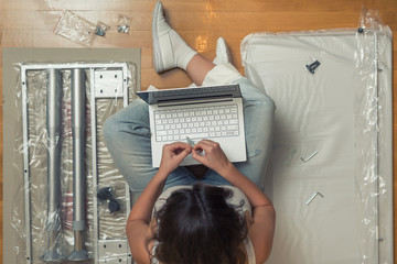Female  is sitting with laptop near parts of furniture for self assembly. Top view