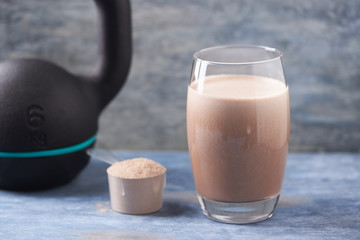 Glass of Chocolate Protein Shake with milk and banana, Whey protein in scoop and black sporting kettlebell in background. Sport nutrition. Rustic wooden background. Copy space. 
