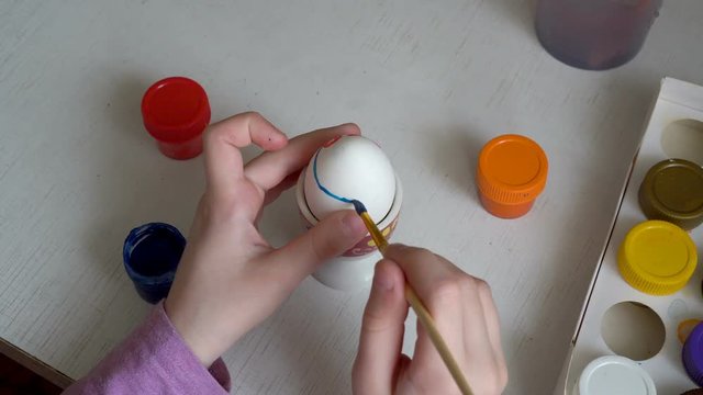 A little girl draws on a white Easter egg with paints and a brush. Close up. The view from the top. 4K. 25 fps