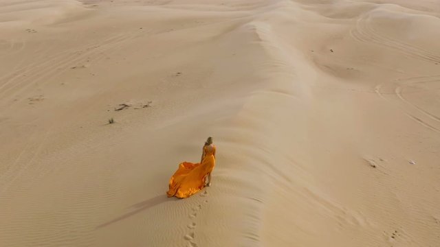 Woman walks in desert on sand-dune. Blonde girl walking on barkhan in hot desert under sun, wind ruffles her hair. Aerial view. Air shoot