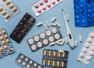 Pink, white, yellow tablets in blisters and thermometer on a blue background top view. Medical pharmacy concept. Traditional medicine.