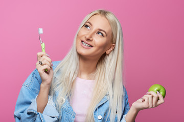Dentist and orthodontist concept. Young blond smiling woman cleaning and brushing teeth with braces using toothbrush. Trying to bite a green apple. Pink background