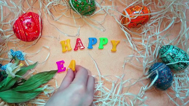 Woman put colourful words Happy Easter on table decorated with easter eggs, blue and white flowers and decorative straw. Top view
