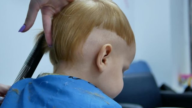 little child boy at hairdresser salon making modern haircut with comb hairbrush and scissors. from back 4k video footage