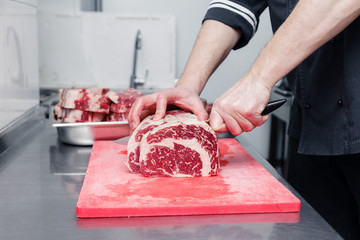 Closeup cook's hand cuts ribeye marbled beef steak with sharp knife on red plastic cutting board on...