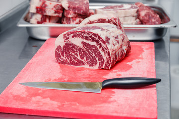 Closeup macro pieces ribeye marbled beef steak with knife on red plastic cutting board on metal table in restaurant kitchen. Concept packing semifinished products, steakhouse, grill meat