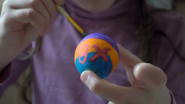 A little girl holds an Easter egg in her hand and paints on it a brush and colored paints, sitting at home at the table. Close up. 4K. 25 fps