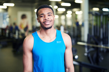 Fototapeta na wymiar Happy young African-american sportsman or coach in blue vest standing in gym in front of camera
