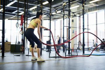 Obraz premium Young active woman with bent knees standing on the floor and exercising with cross training ropes in gym