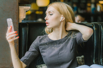 The blonde takes a selfie in the cafe, she has a wireless earpiece in her ear.