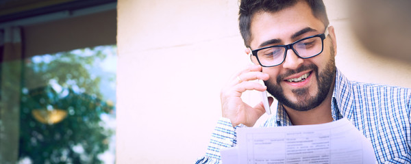 Young bearded businessman in casual clothes and eyeglasses is studying document while talking on the smartphone sitting on the terrace of a summer cafe.