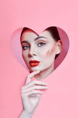 Woman looking in the heart hole, bright beautiful makeup, big eyes and lips, bright lipstick, professional cosmetics and facial care. Bright colored background and a gap slot in the paper heart