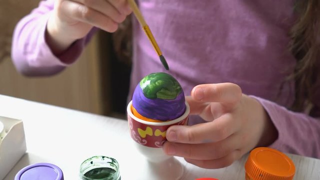A little girl paints an Easter egg with colorful colors sitting at home at the table. Close up. 4K. 25 fps