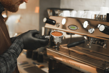 Fototapeta na wymiar Close up photo of male barista holding portafilter at the coffee machine at the coffee shop. Barista's hands holding a holder with roasted coffee at the cafe. Preparing coffee concept.