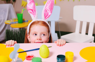 Obraz na płótnie Canvas cheerful girl a child with red hair with rabbit ears sits under the table and hunts the Easter bunny. Easter traditions.