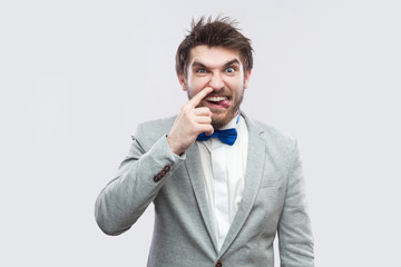 Portrait of funny handsome bearded man in casual grey suit and blue bow tie standing and looking at camera with tongue out and drilling nose. indoor studio shot, isolated on light grey background.