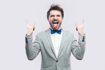 Portrait of funny crazy handsome bearded man in casual grey suit and blue bow tie standing with rock gesture, closed eyes and tongue out. indoor studio shot, isolated on light grey background. - obrazy, fototapety, plakaty