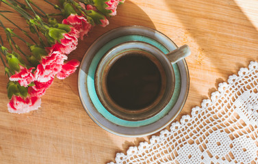 Close up view of a blue vintage cup of coffee, pink carnations and white doily on wooden background illuminated by sun rays coming from a window. Good morning coffee or hello spring