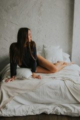 Beautiful tender girl in black lingerie and jumper is sitting on the bed. A young woman basks in bed with white linens. The girl woke up and enjoys a good morning.