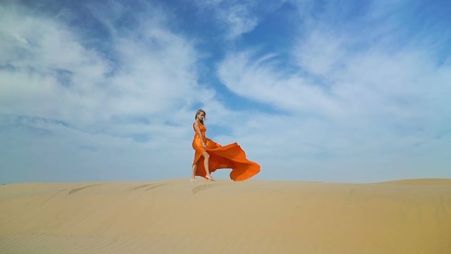 Girl walks in desert on barkhan. Woman in dress walking on sand-dune in hot desert under sun, wind ruffles her hair