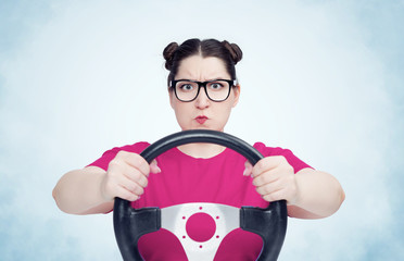 Serious young girl in glasses and t-shirt with steering wheel on light blue smoke background, car driver concept