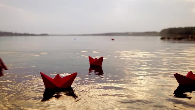 Woman's hand is putting red paper boat on the water to others swimming at sunset. Origami ships on waves on river in the evening