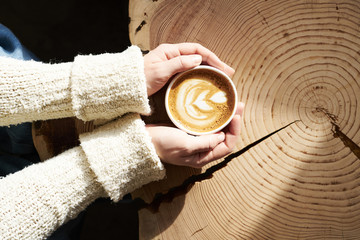 Closeup view woman hands holding cup of coffee at wooden slab table, top view, sunny cafe, warm atmosphere.