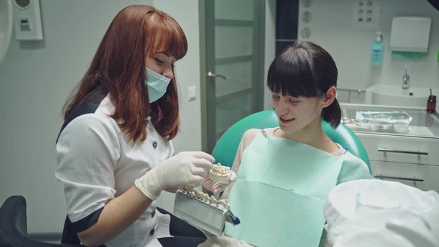 Friendly dentist showing jaw layout to a patient girl sitting in the stomatology clinic. Professional dentist explaining to female patient correct tooth brushing on the dental model