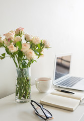 Close up of pale pink roses in glass with home office desk in background including, laptop, notebook, pen and glasses (selective focus)