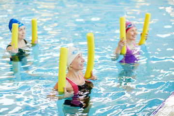 Side view at group of active senior women exercising in swimming pool, holding pool noodles and smiling, copy space - Powered by Adobe