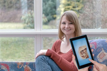 Fototapeta premium Smiling young woman is sitting at home and another female hands are showing space shot of the unborn child in the tablet screen.