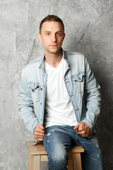 Young handsome guy posing in denim clothes sitting on a chair against the background of a concrete wall