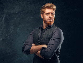 Handsome elegantly dressed redhead man posing with his arms crossed in studio against a dark textured wall