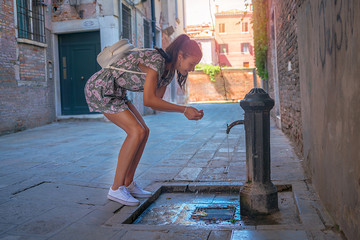 hipster teenager girl drinks water and washes on a hot day on a city street near tap with drinking water