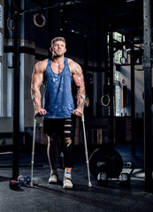 Fototapeta na wymiar Muscular bodybuilder standing with crutches and bandage on a leg in a gym. Injury recovery, sport, motivation