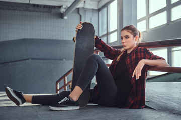 A beautiful girl wearing a checkered shirt holding a skateboard while sitting next to a grind rail in skatepark indoors.