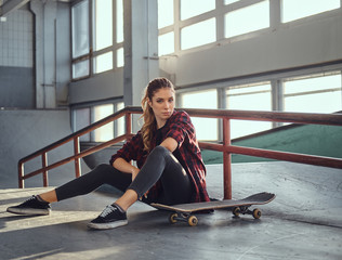 A beautiful girl wearing a checkered shirt holding a skateboard while sitting next to a grind rail...