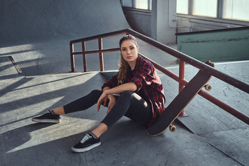 A beautiful girl wearing a checkered shirt holding a skateboard while sitting next to a grind rail...