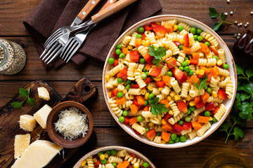 Italian pasta salad with fusilli, corn, boiled carrot, fresh green peas and sweet pepper on wooden rustic background. Top view
