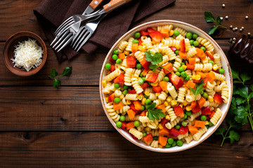 Italian pasta salad with fusilli, corn, boiled carrot, fresh green peas and sweet pepper on wooden rustic background. Top view