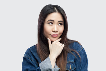 Portrait of dreamy smiley beautiful brunette asian young woman in casual blue denim jacket with makeup standing touching her chin and pondering. indoor studio shot, isolated on light grey background.