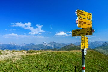 Gelber Wanderwegweiser auf einer gürnen Bergspitze im Engadin in der Schweiz bei blauem Himmel und wenigen weissen Wolken