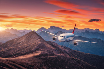 Airplane is flying over mountains at colorful sunset in summer. Landscape with passenger airplane, hills, orange sky with red clouds. White aircraft. Business travel. Commercial plane. Aerial view
