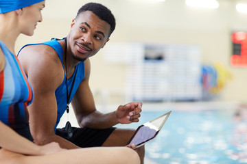 Portrait of handsome African-American fitness coach talking to client in swimming pool, copy space