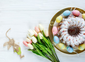 Happy Easter! Golden tray with plate wit cake and hand painted colorful eggs, tulips on white wooden table. Close up. Decoration for Easter, festive background.