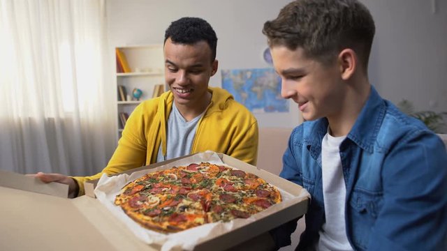 Two multiracial friends opening box with pizza for student party, food delivery