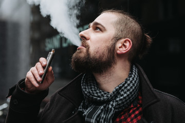 Men with beard hold and smoke his electronic cigarette outdoor in black, grey backgraund