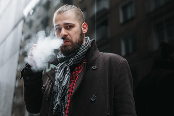 Fototapeta na wymiar Men with beard hold and smoke his electronic cigarette outdoor in black, grey backgraund